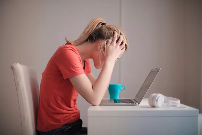 Stresul și sănătatea. Tânără copleșită de stres, cu capul în mâini, în fața laptopului